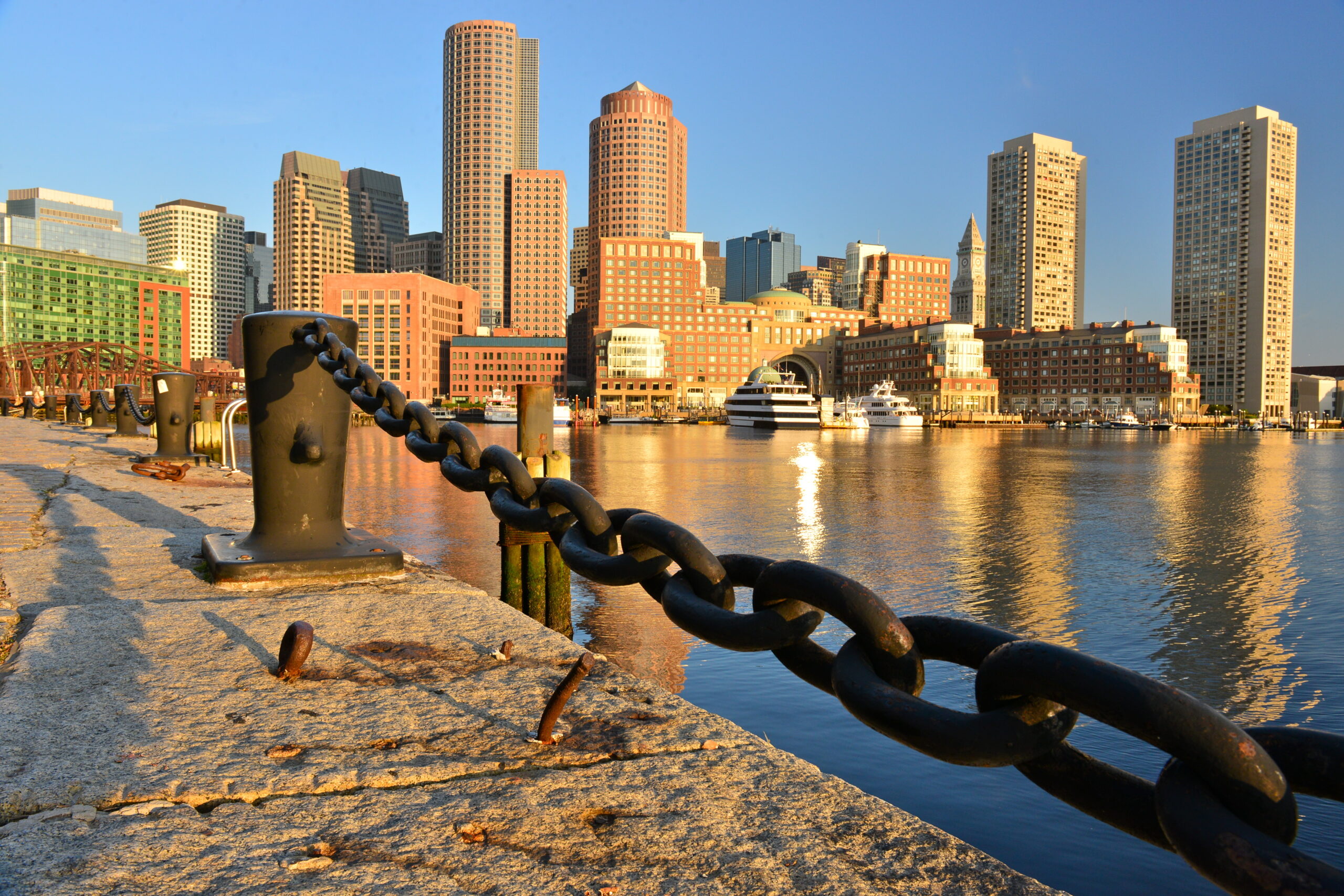 View of Boston Harbor with waterfront chains and the skyline of the Financial District reflected in the water during daylight.