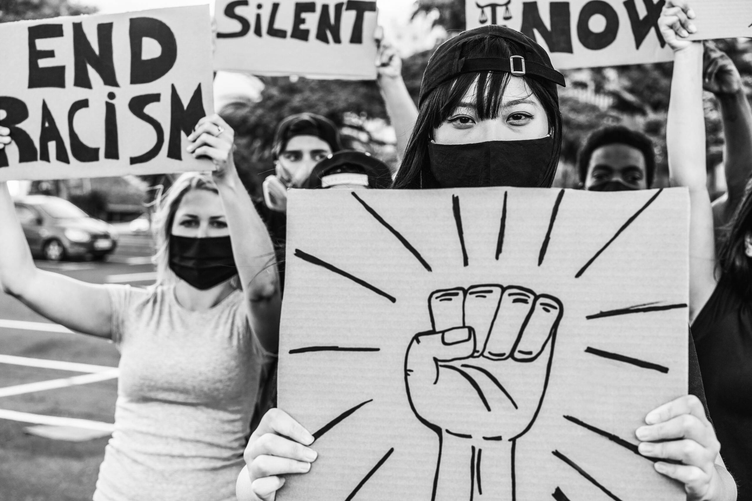 Black-and-white photo of Asian American protesters at a demonstration, including a masked woman holding a protest sign with a raised fist symbol.