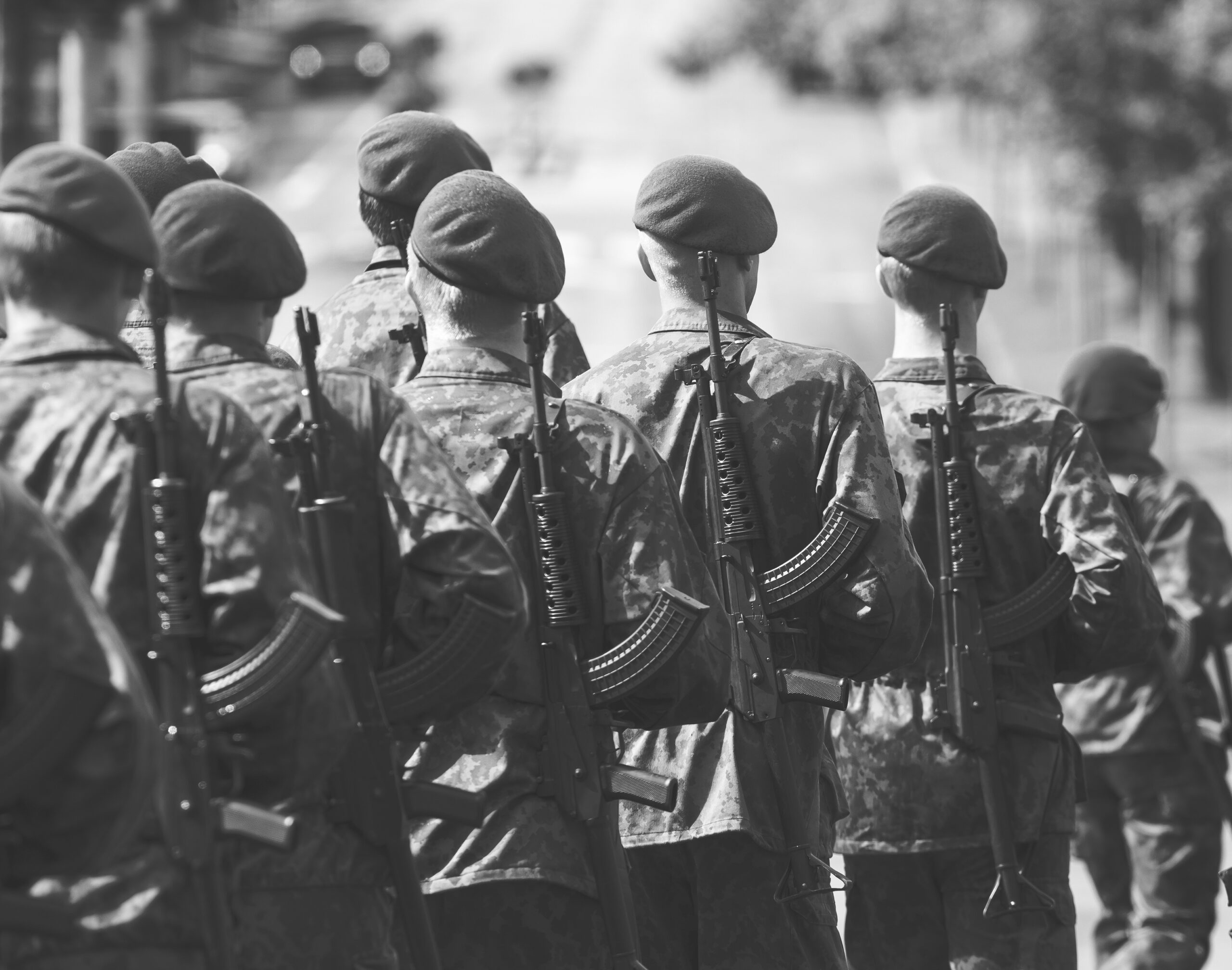 Black-and-white image of soldiers marching in formation during a military parade, viewed from behind with rifles over their shoulders.