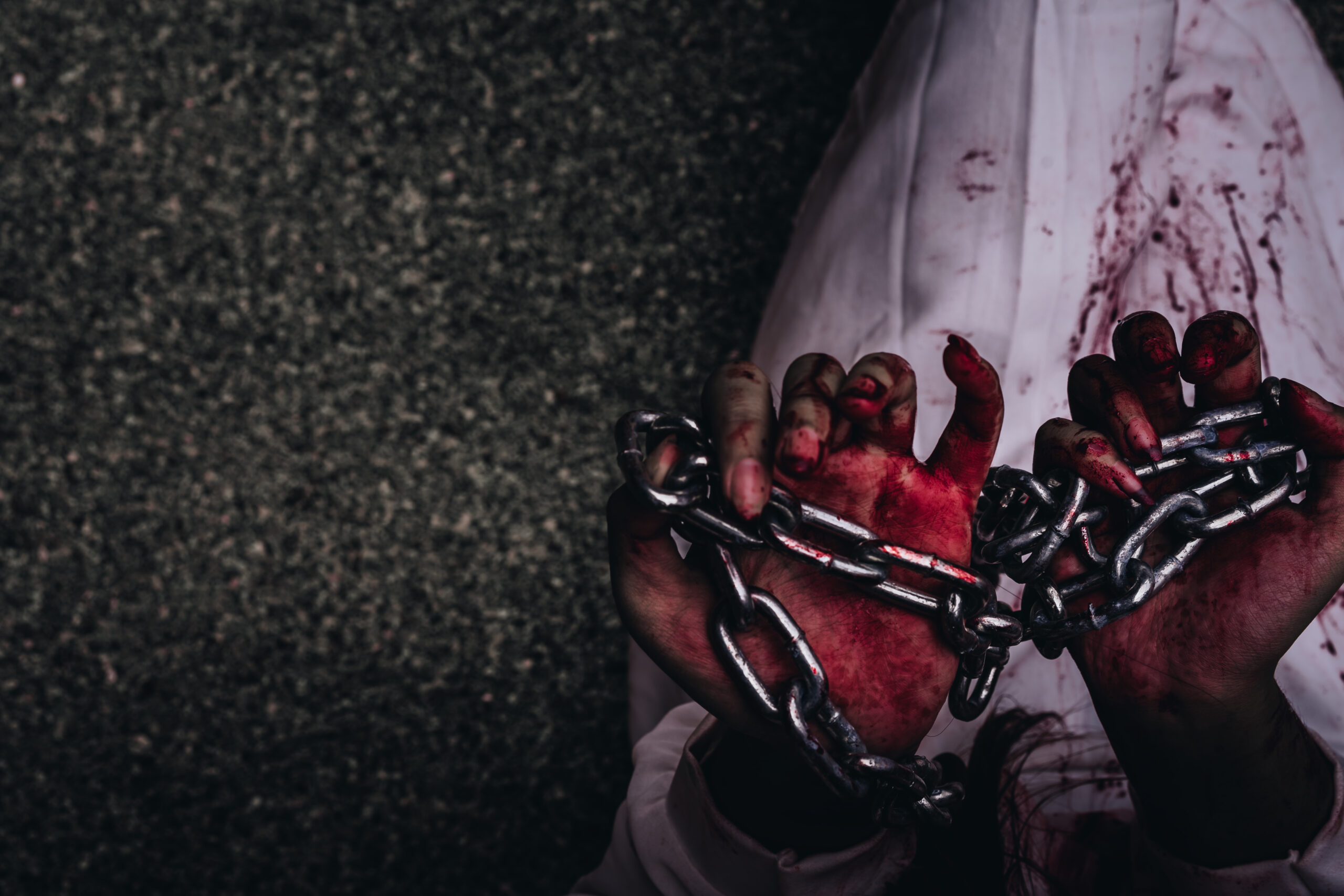 Close-up image of bloodied hands gripping metal chains in a dark setting, symbolizing violence, imprisonment, torture, or human suffering.