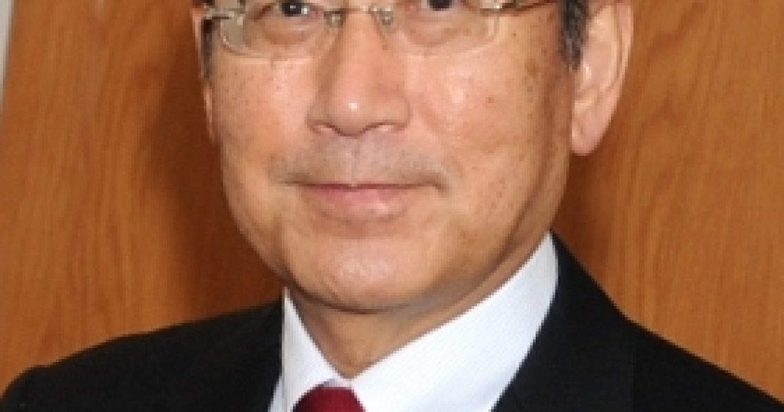 Portrait of Tadatoshi Akiba wearing glasses and a suit with a red tie against a wooden background.