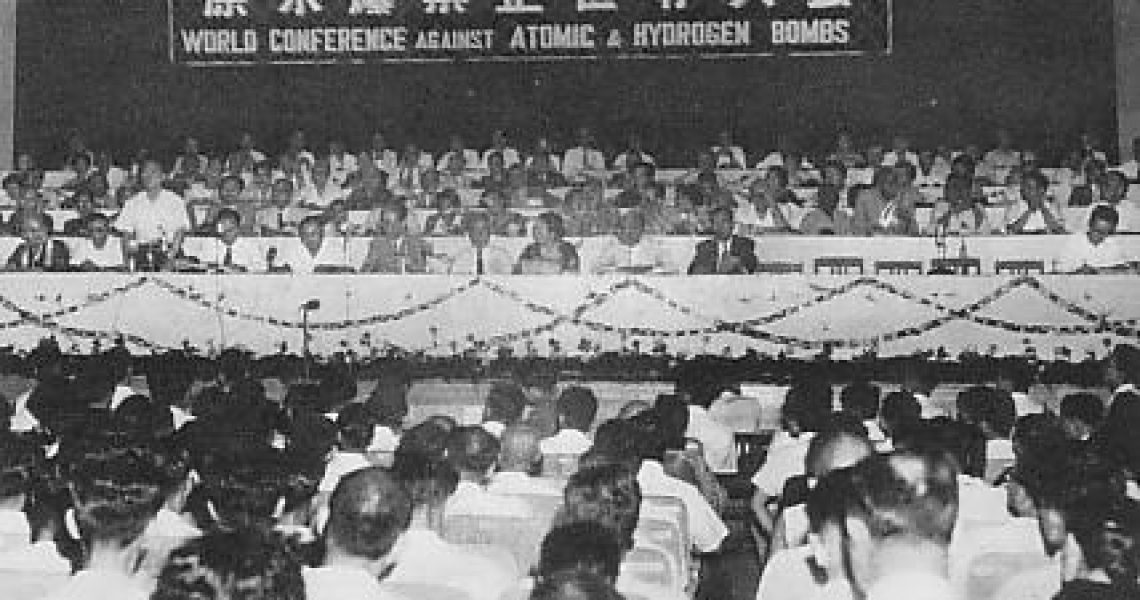 Historic black-and-white photograph of the First World Conference Against Atomic and Hydrogen Bombs, showing a large audience beneath banners advocating nuclear disarmament.