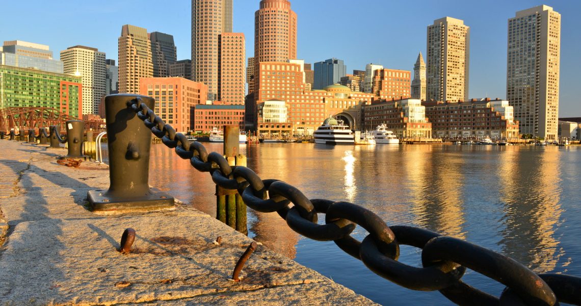 View of Boston Harbor with waterfront chains and the skyline of the Financial District reflected in the water during daylight.