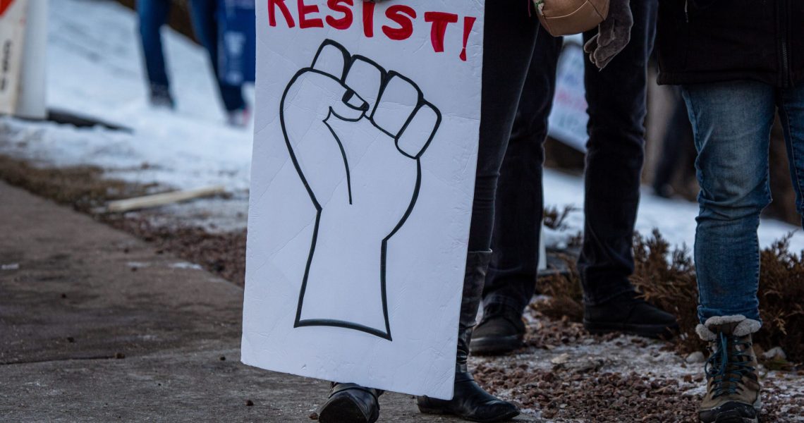 Close-up of a protester holding a sign with a raised fist illustration and the word “RESIST!” written in red during a public demonstration.