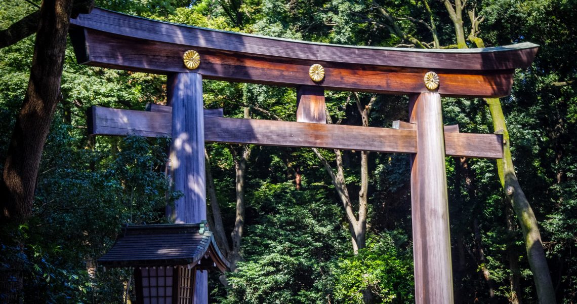 Traditional Japanese Shinto shrine gate surrounded by trees and forest greenery, featuring wooden beams and decorative gold accents.