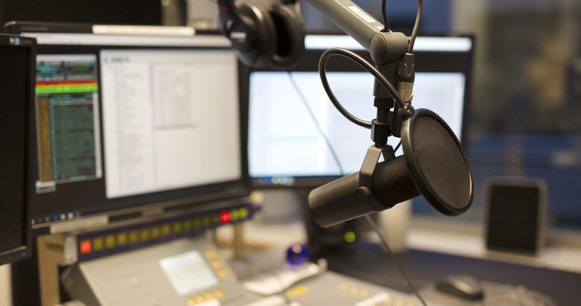 Close-up of a professional microphone in a modern radio broadcasting studio with computer monitors and audio equipment in the background.