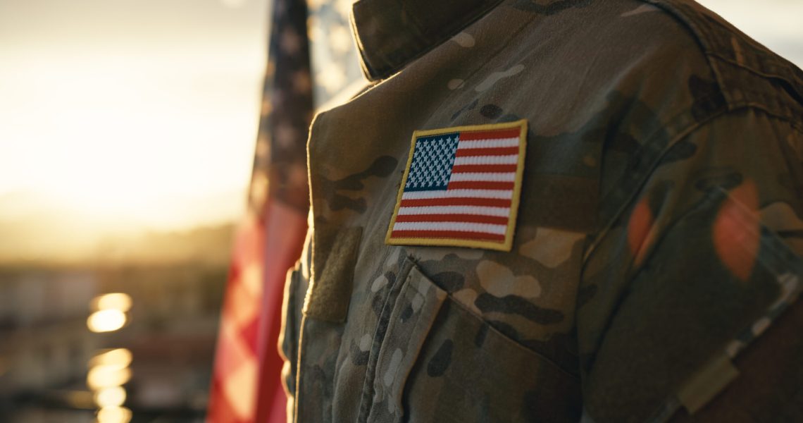 Close-up of a U.S. military uniform featuring an American flag patch, with a blurred flag and warm sunlight in the background.