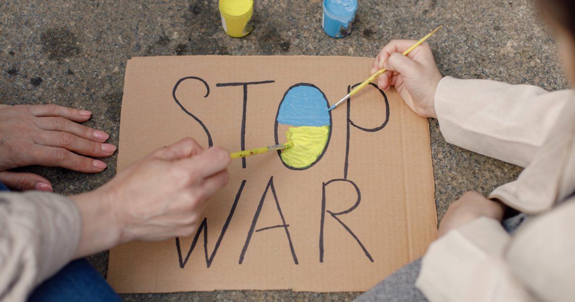 Cropped photo of a poster with phrase Stop War on it. Woman and girl painting it in blue and yellow colors. Poster for supporting Ukraine.