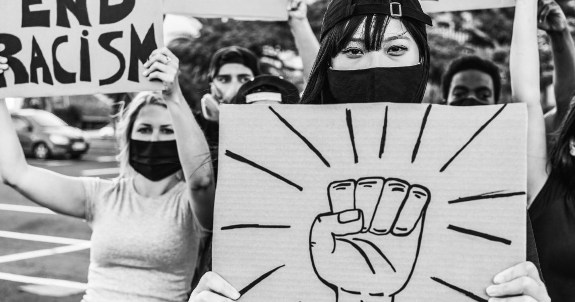 Black-and-white photo of Asian American protesters at a demonstration, including a masked woman holding a protest sign with a raised fist symbol.
