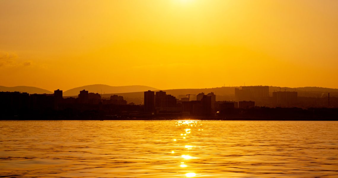 Sea panorama of the city at sunset, silhouettes of the coast and buildings against the backdrop of the orange setting sun reflected in the water