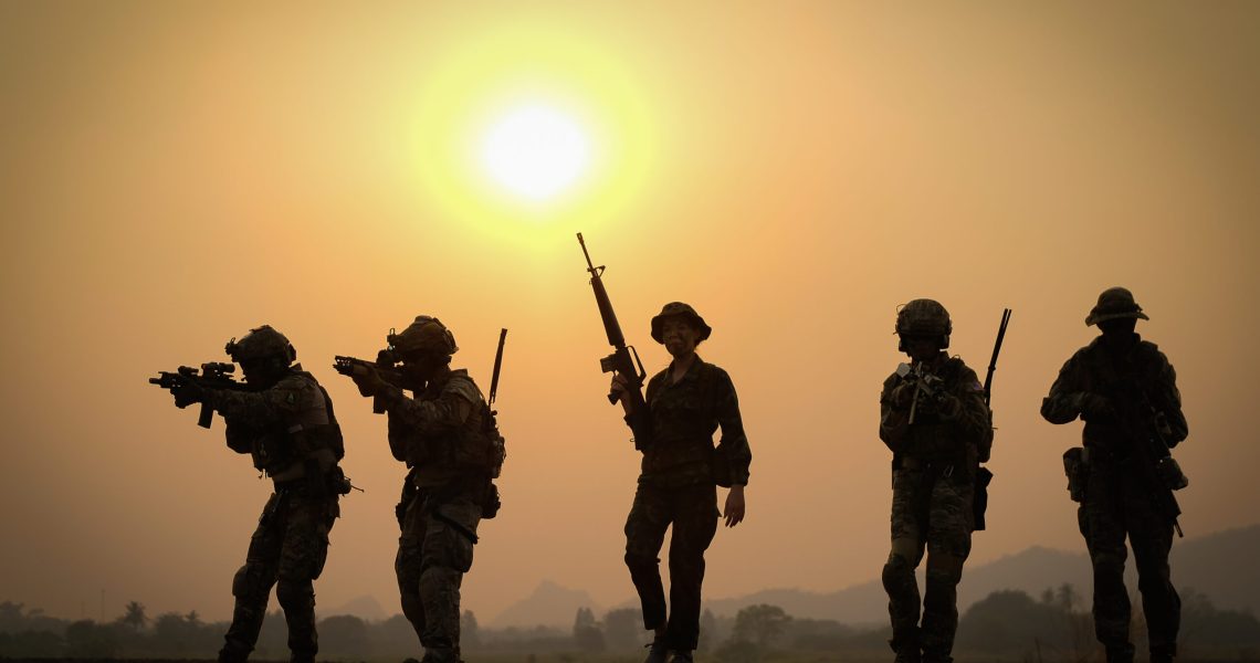 Silhouettes of armed soldiers standing against a sunset sky, with the sun glowing behind them and weapons visible in profile.